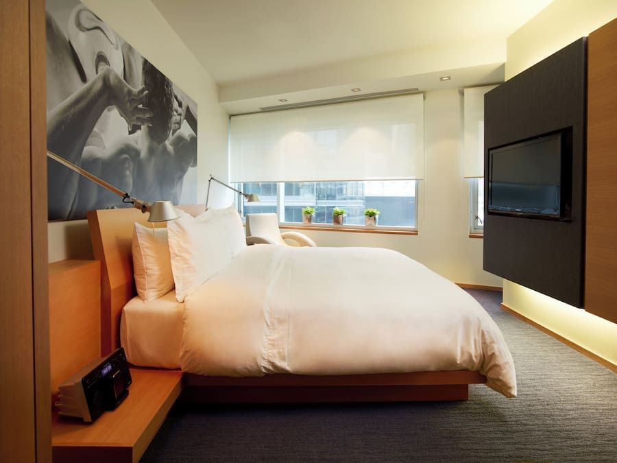 A modern hotel room with a large bed, a bedside table with a lamp, a TV mounted on the wall, and a window with a view of buildings.