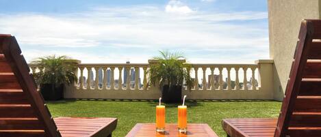 A patio with two wooden lounge chairs and a table, two drinks on the table, and a view of a city skyline through a white fence.