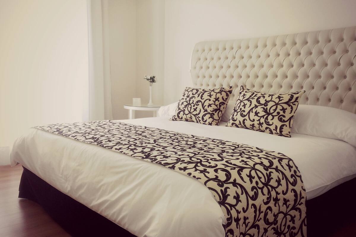 A neatly made bed with a patterned bedspread and two decorative pillows, a small bedside table with a vase of flowers, and a tufted headboard.