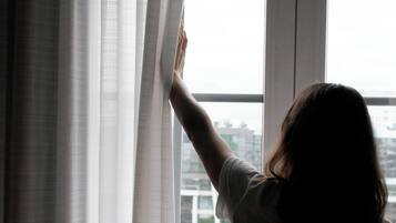 A person standing by a window with sheer curtains, looking outside.