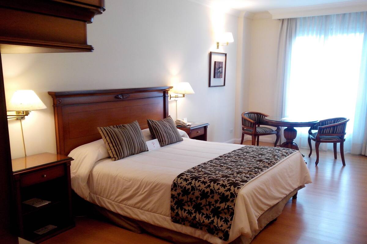 A hotel room with a large bed, two bedside tables with lamps, a small table with chairs, and a window with curtains.