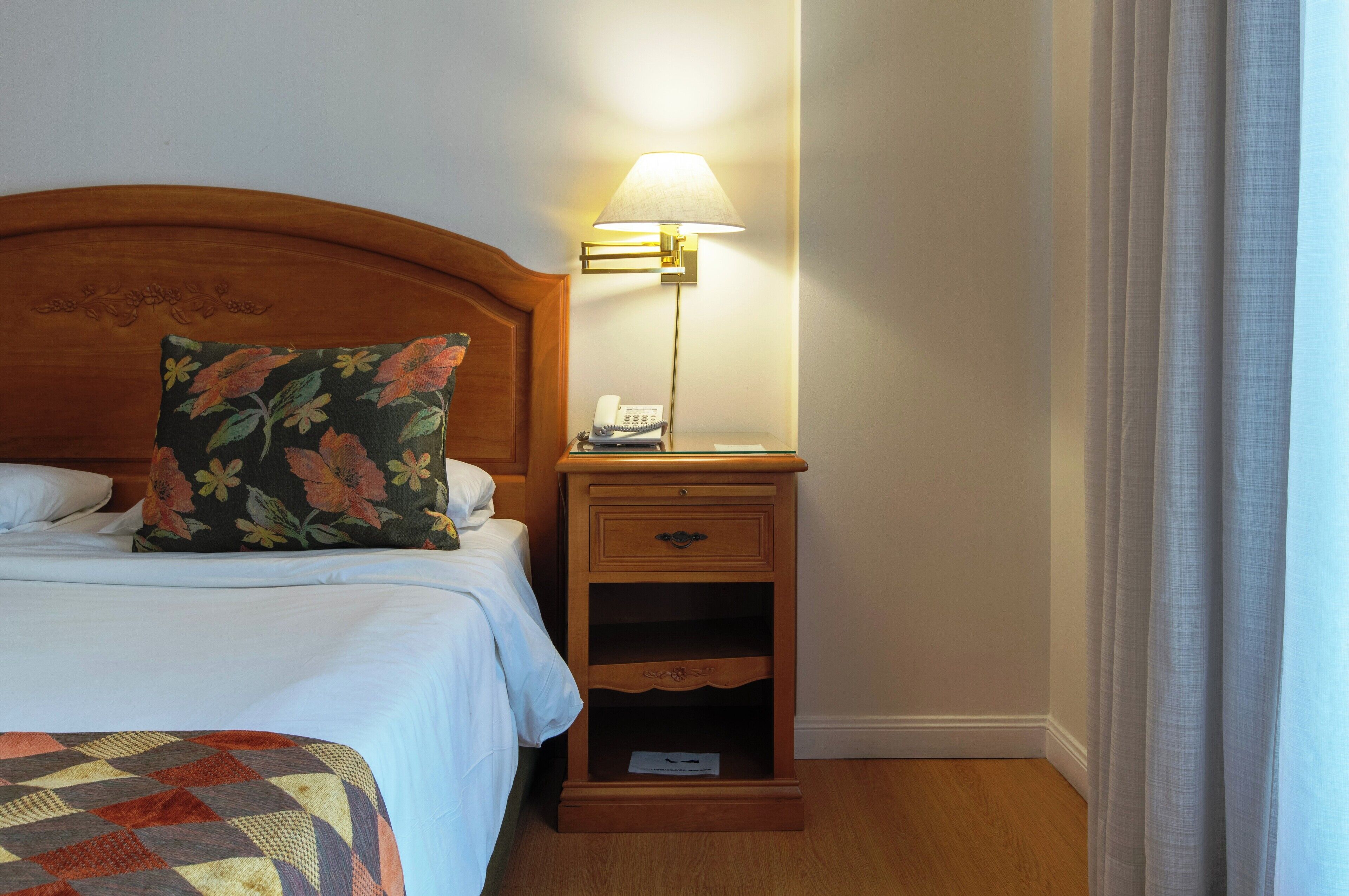 A hotel room with a wooden headboard, a floral pillow, a bedside table with a lamp, and a quilt on the bed.