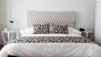 A neatly made bed with a tufted headboard, two patterned pillows, and a white bedspread. There are two bedside tables, one with a vase and a sign, and the other with a phone and a lamp.