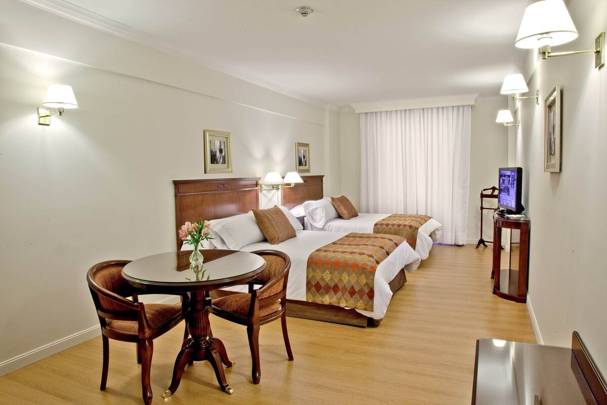 A hotel room with a wooden bed, two bedside tables, a round dining table with chairs, a TV on a stand, and a large window with curtains.
