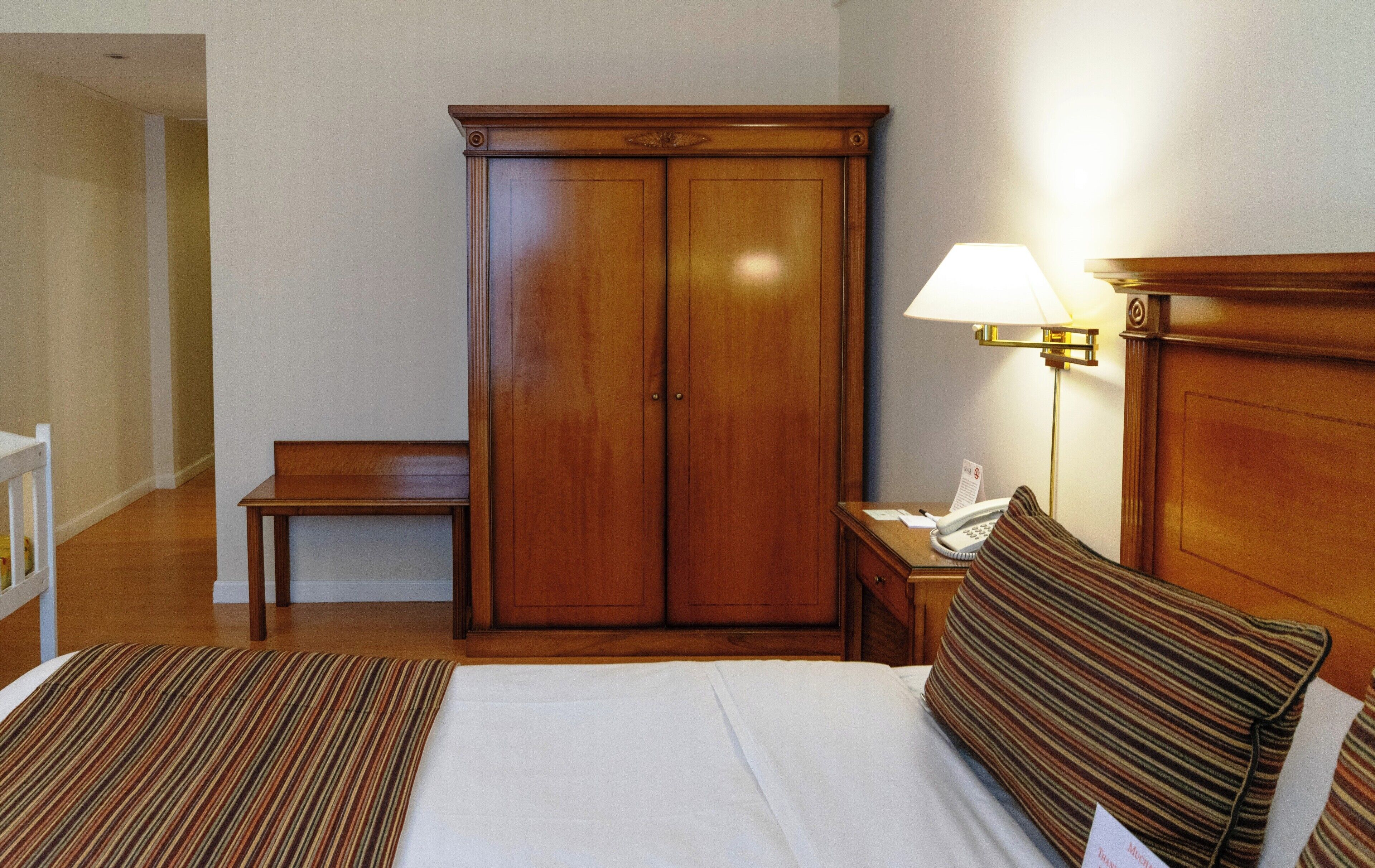 A hotel room with a bed, a bedside table with a lamp, a wooden wardrobe, and a small bench.