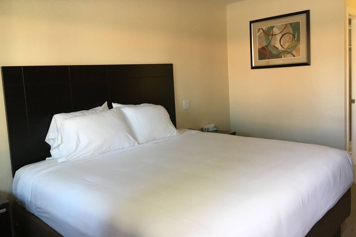A large bed with white linens, a dark headboard, and a framed abstract painting on the wall.