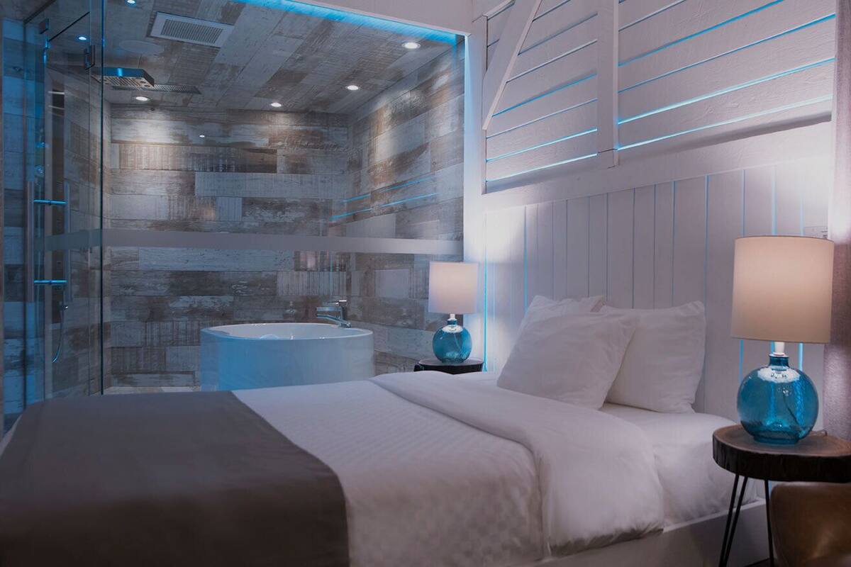 Una habitación de hotel moderna con una cama grande, un box de ducha con paredes de vidrio, una bañera redonda y una mesita de noche con lámpara.