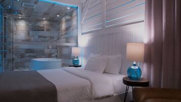 A modern hotel room with a large bed, a glass-enclosed shower, a round bathtub, and a bedside table with a lamp.