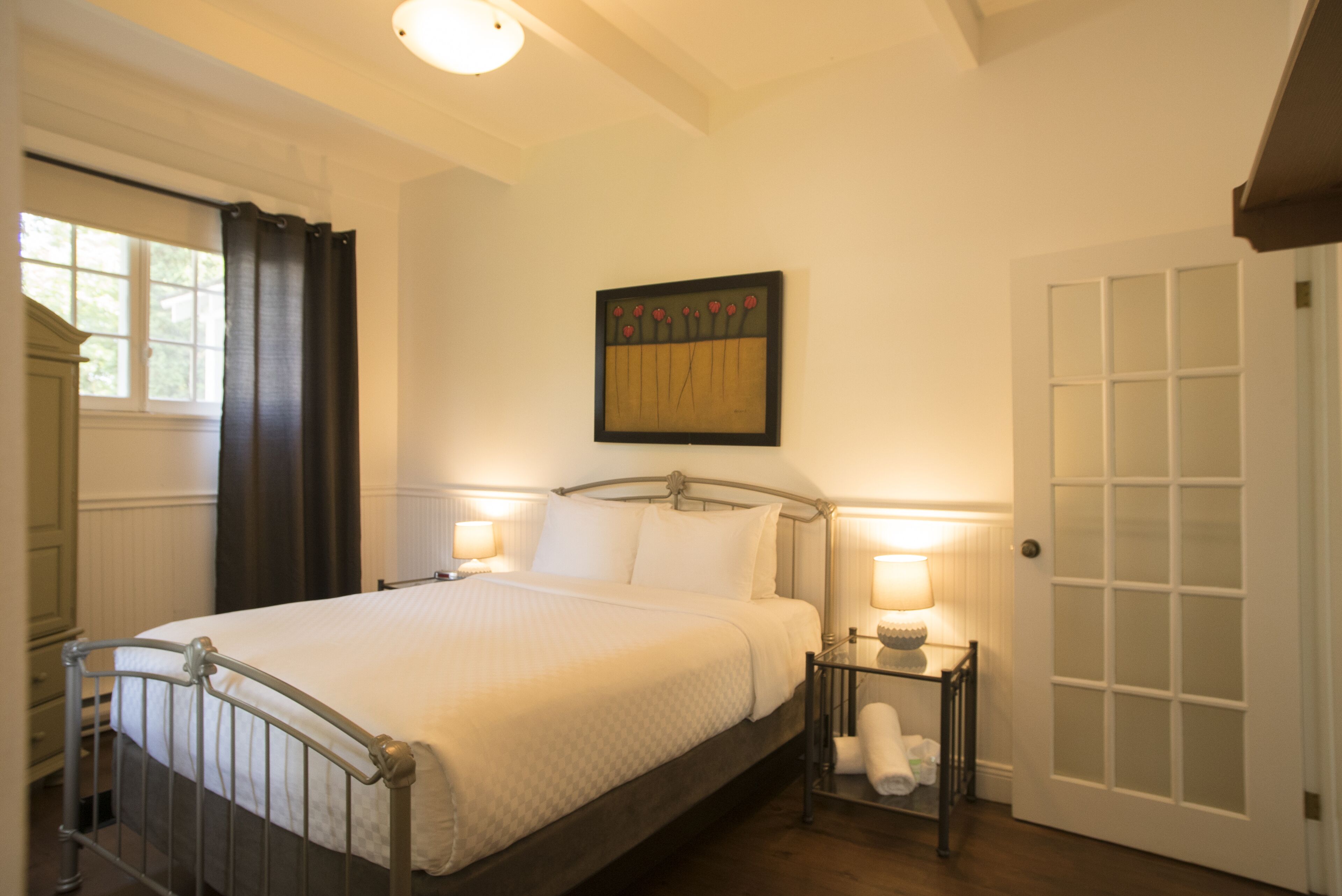 Ein ordentlich eingerichtetes Schlafzimmer mit Bett, Nachttischlampen, einem gerahmten Kunstwerk und einer Tür mit Glasfeldern.