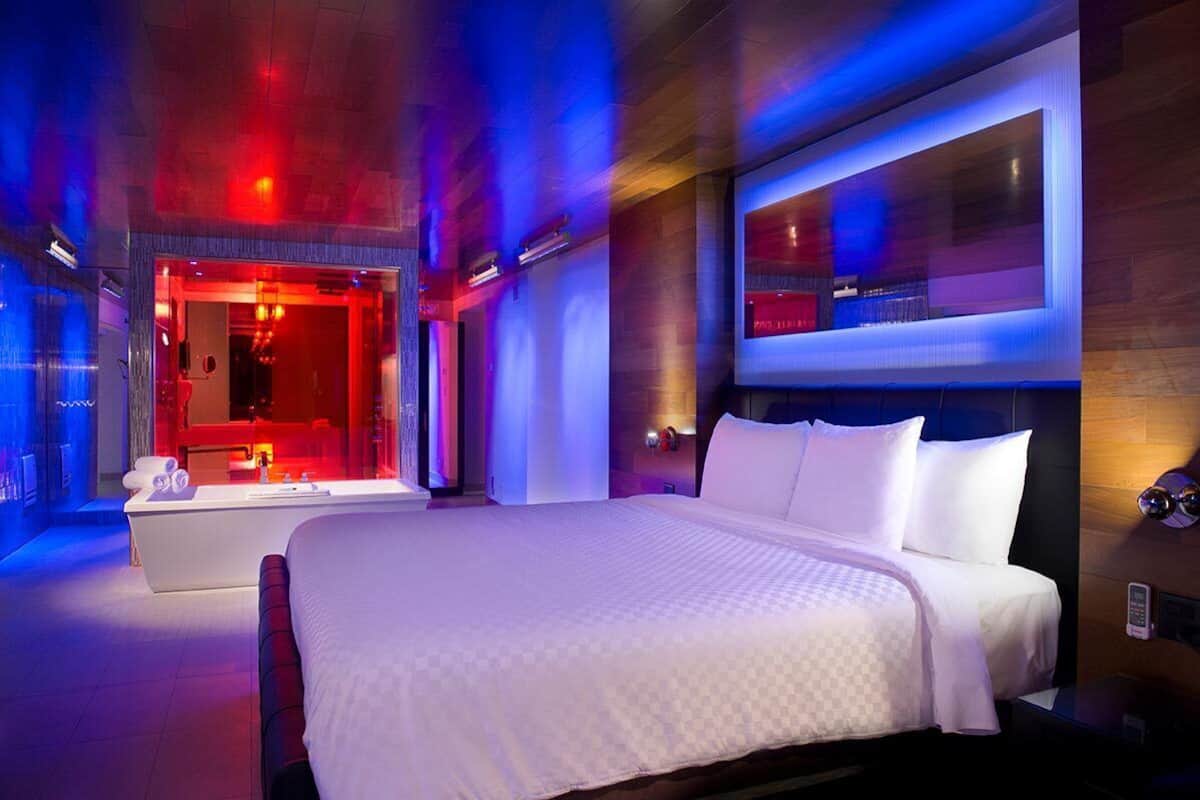 Una habitación de hotel moderna con una cama grande, una zona de estar separada con tocador iluminado en rojo, y un ventanal con vistas.