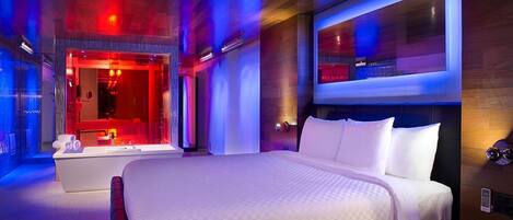 A modern hotel room with a large bed, a separate seating area with a red-lit vanity, and a large window with a view.