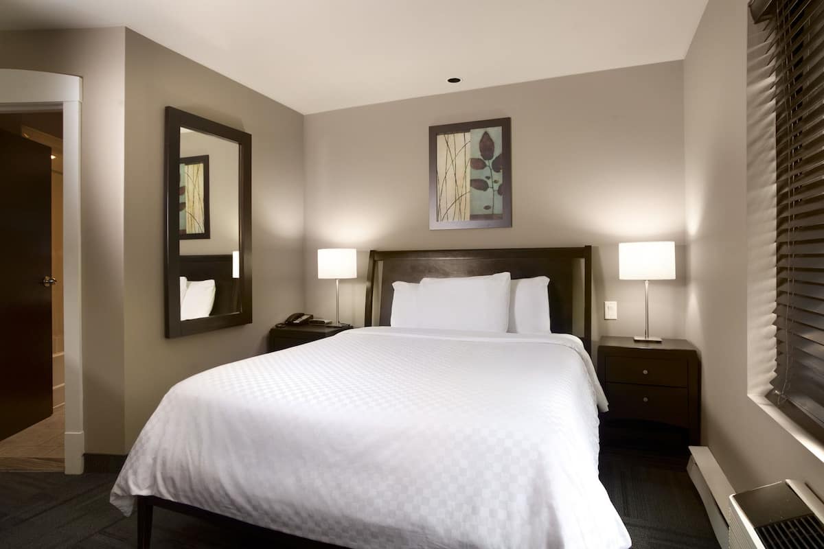 Un cuarto de hotel con una cama grande, dos mesitas de noche con lámparas, un tocador, un espejo en la pared y un cuadro sobre la cama.