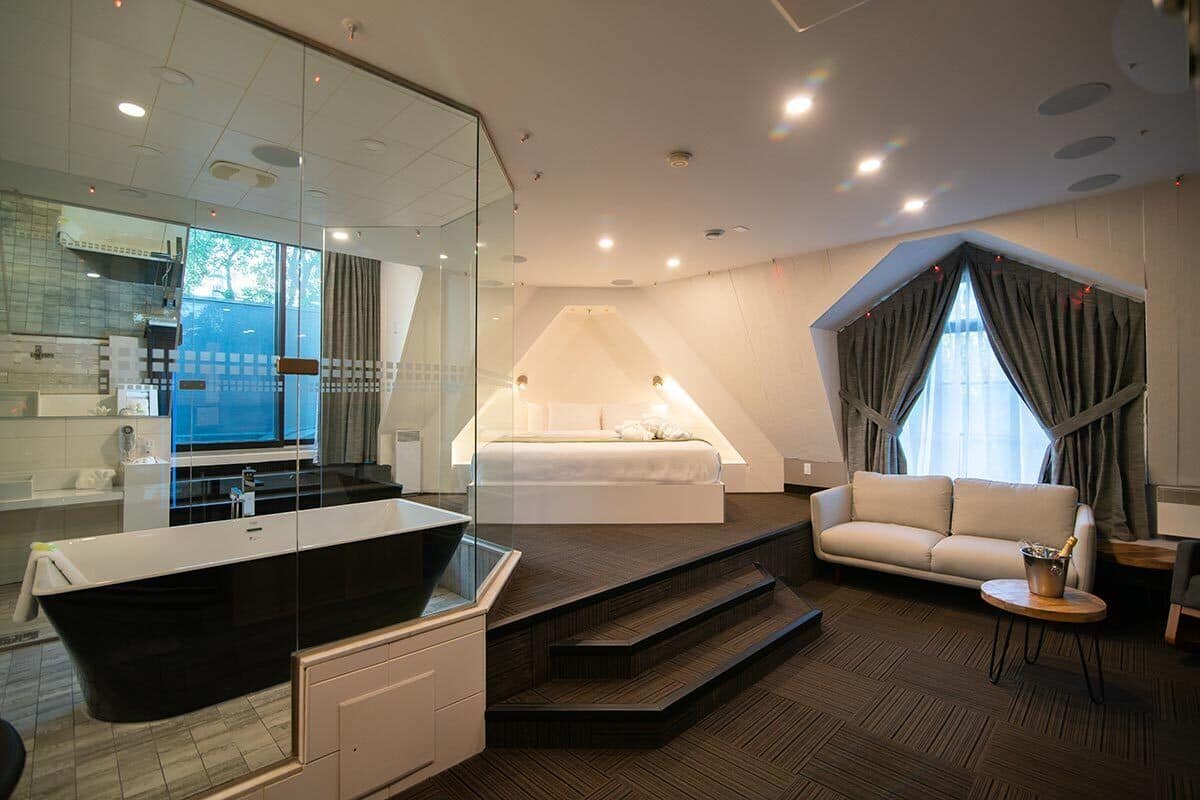 Una habitación de hotel moderna con una bañera negra grande, un dormitorio separado con una cama, un sofá y una mesita con una botella de champán.