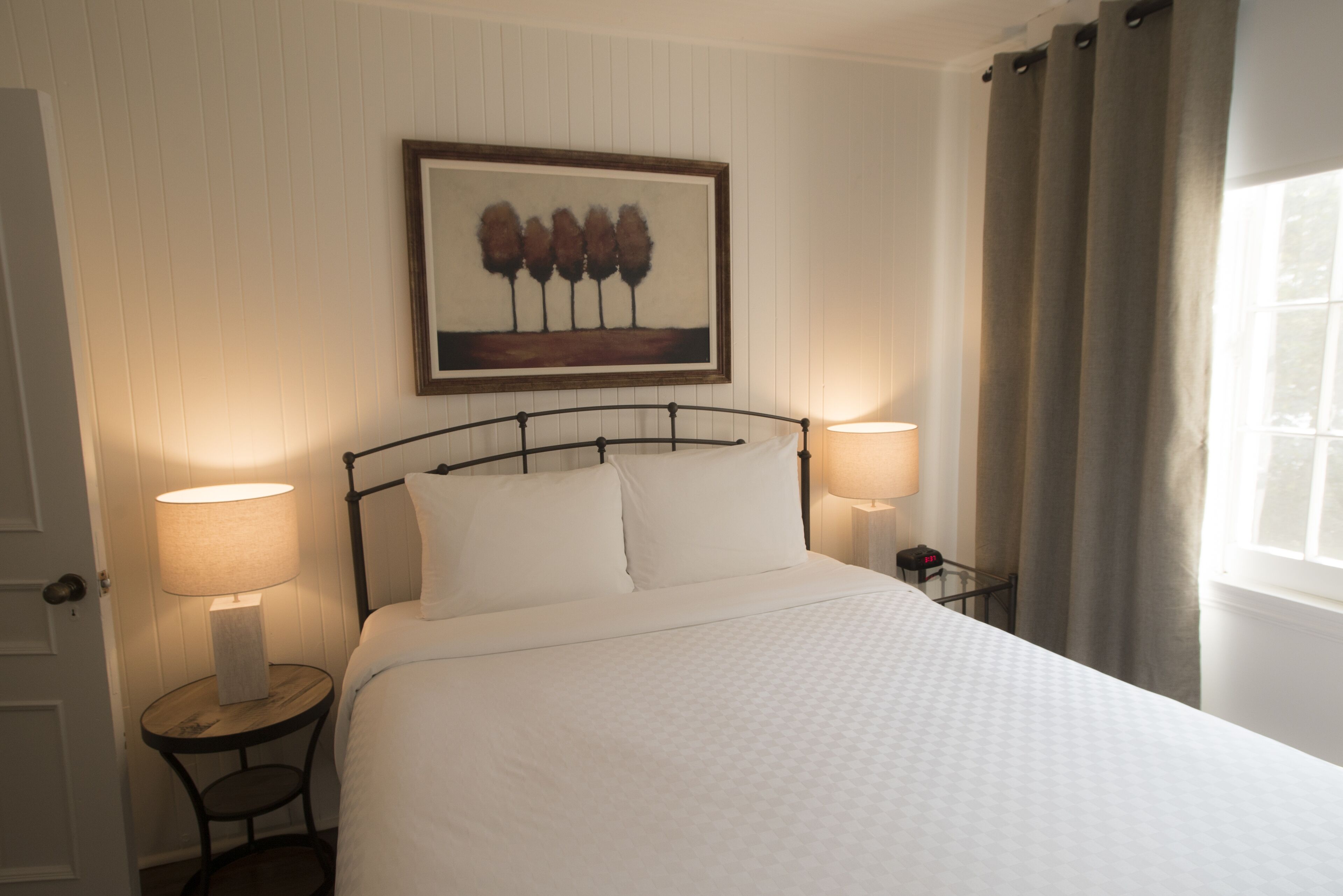 A neatly made bed with white linens, two bedside lamps, a framed artwork above the bed, and a window with curtains.