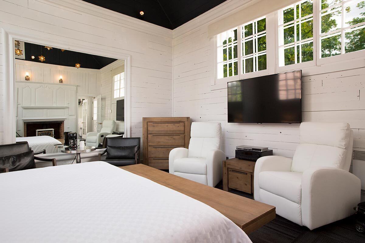 A modern hotel room with a large bed, two white armchairs, a wooden nightstand, a flat-screen TV mounted on the wall, and a fireplace.