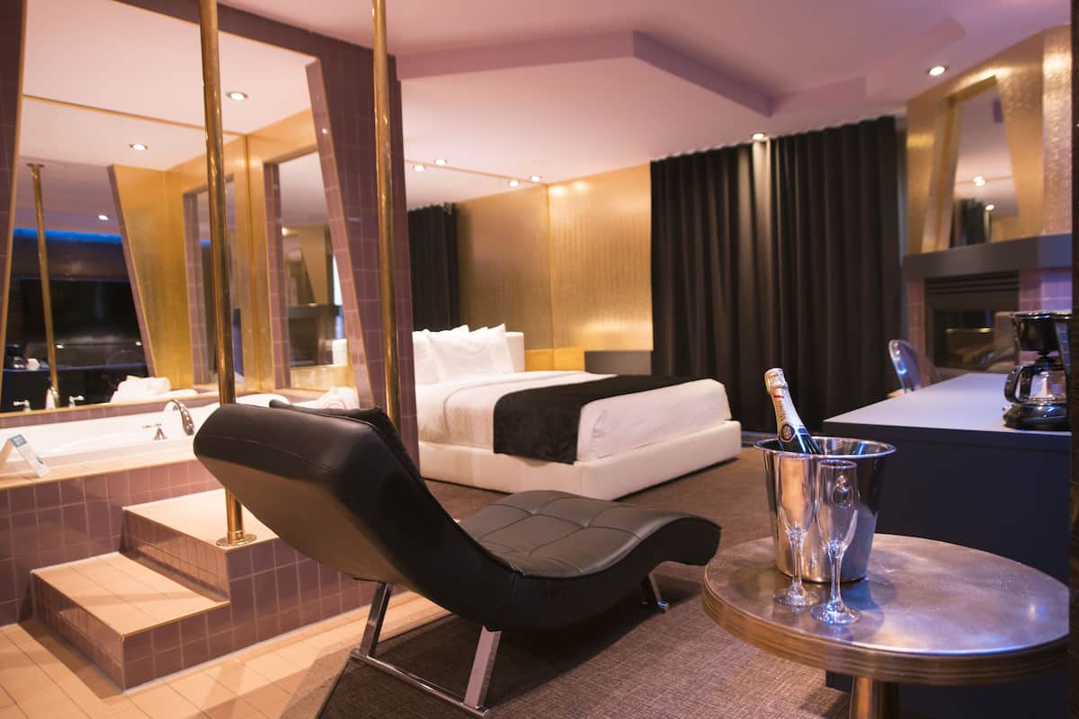 Una habitación de hotel moderna con una cama grande, una zona de estar con una silla, una mesita con una botella y vasos, una cafetera y un espejo grande.