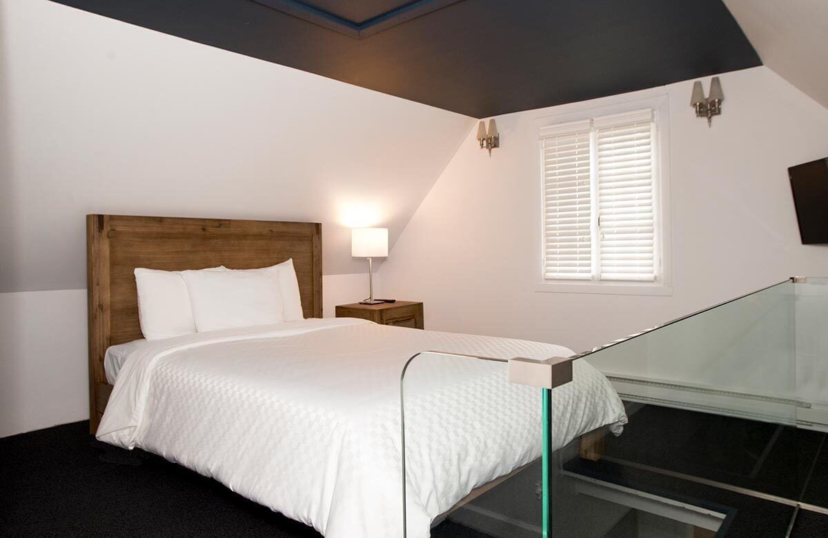 Een slaapkamer met een houten hoofdbord, een bedlampje, een raam met zonwering en een glazen hekwerk.