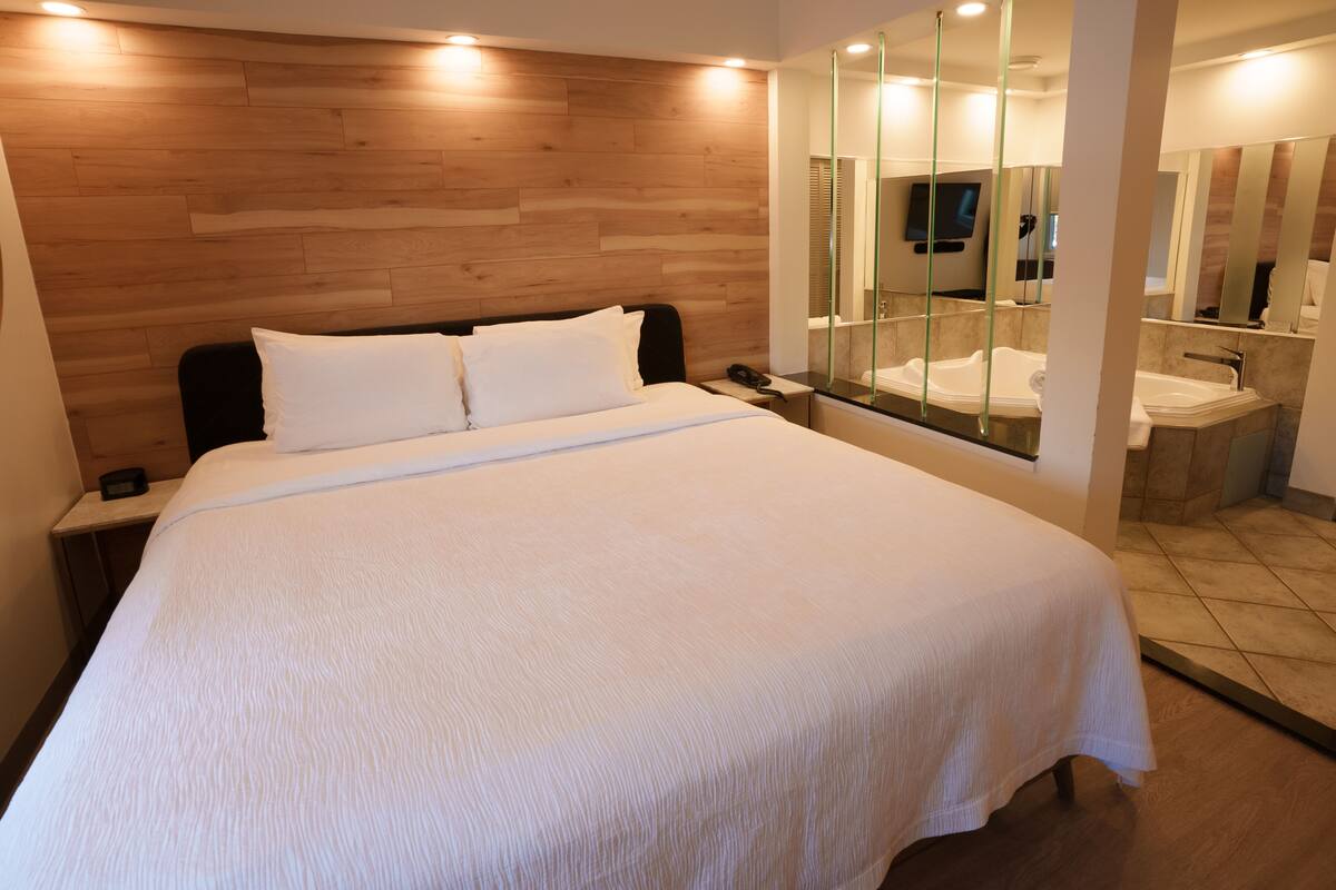 Habitación de hotel con una cama grande, cabecera de madera, mesita de noche con teléfono, y un baño con un espejo grande y dos lavamanos.