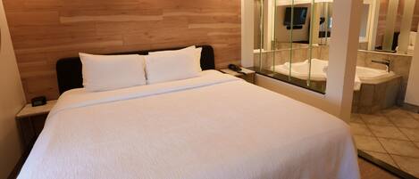 Habitación de hotel con una cama grande, cabecera de madera, mesita de noche con teléfono, y un baño con un espejo grande y dos lavamanos.