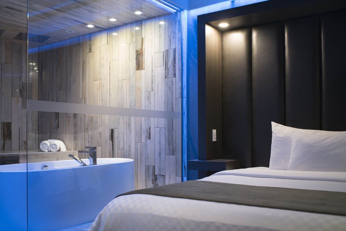 Una habitación de hotel moderna con una cama grande, una bañera acristalada y un cabecero de tonos oscuros.