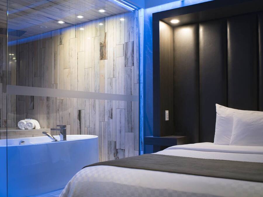Une chambre d’hôtel moderne dotée d’un grand lit, d’une baignoire entourée de parois en verre et d’une tête de lit aux tons sombres.