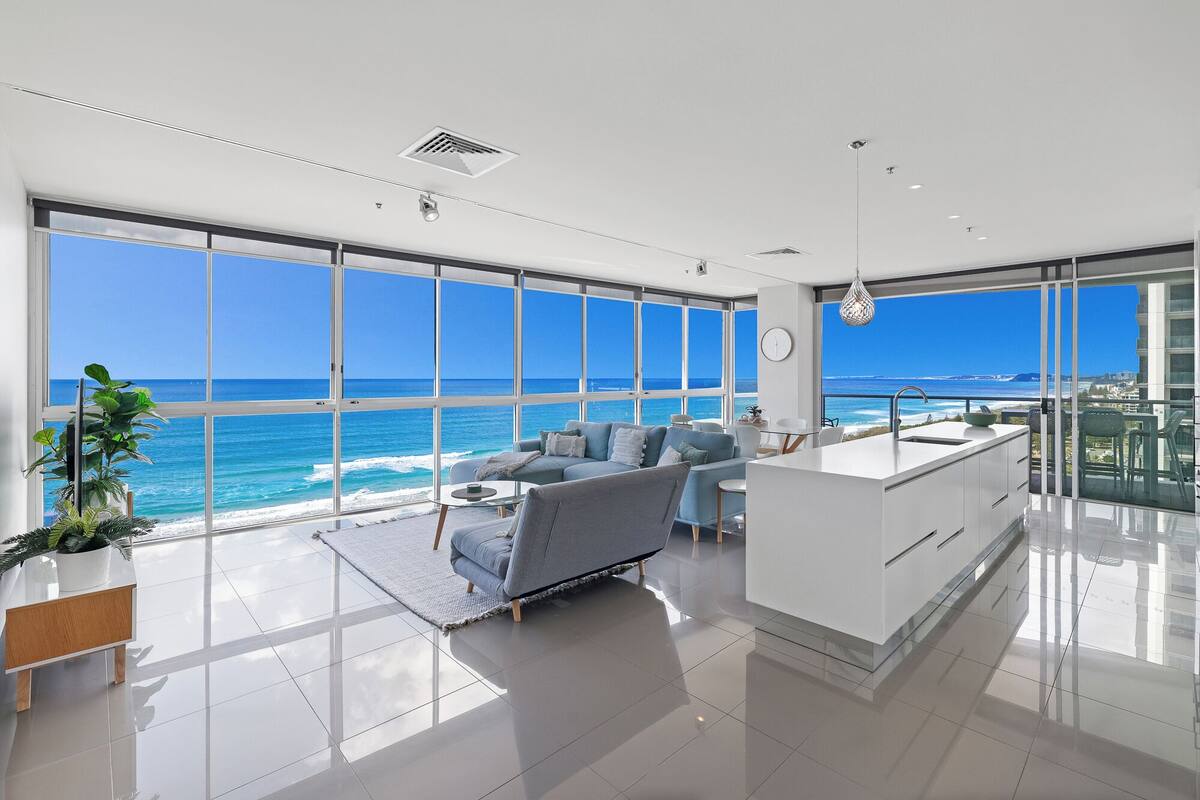 A modern living room with a large window overlooking the ocean, a grey sofa, a small coffee table, and a kitchen area with a sink and island.