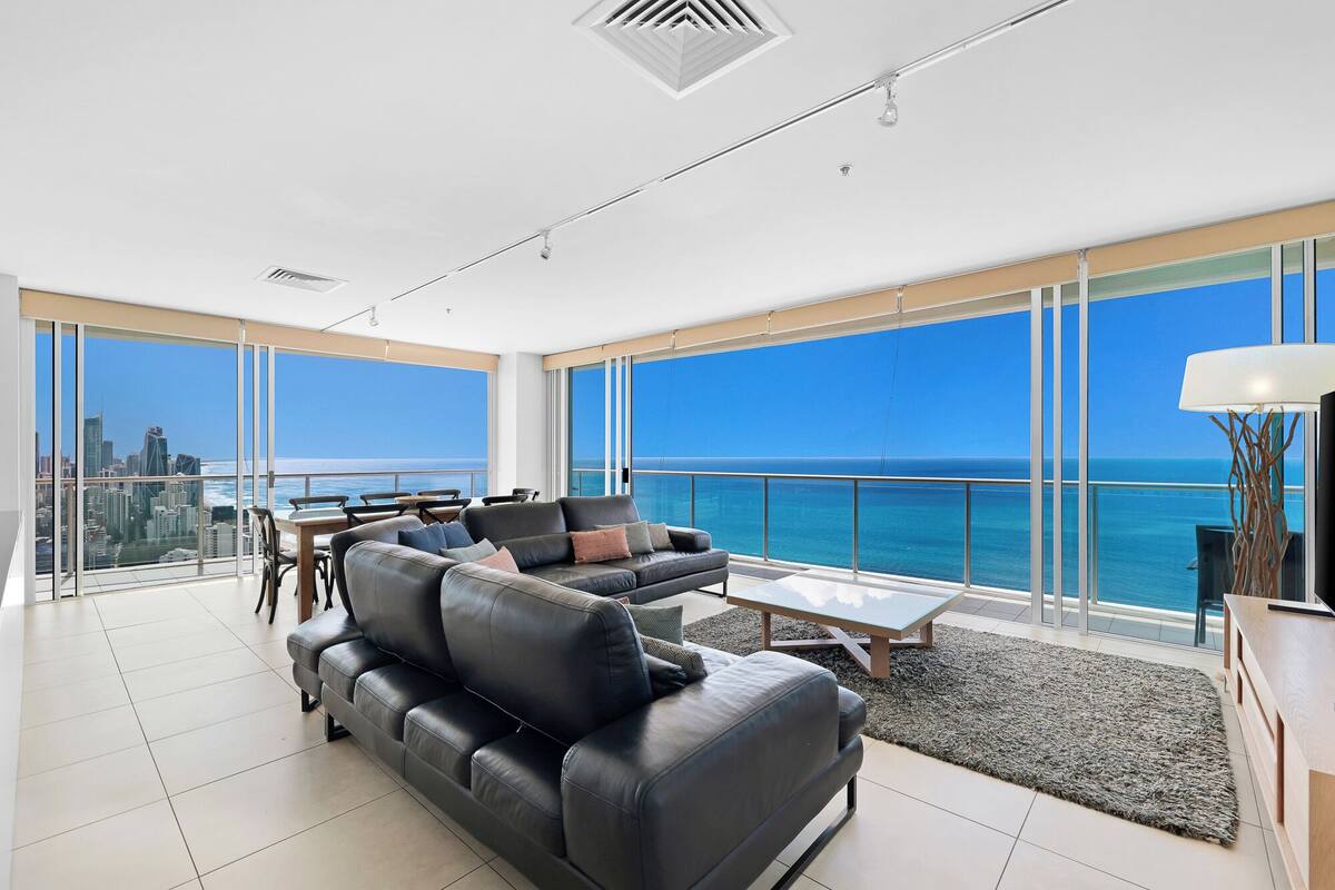 A spacious living room with a large black leather sofa, a coffee table, and a view of the ocean and city skyline through floor-to-ceiling windows.