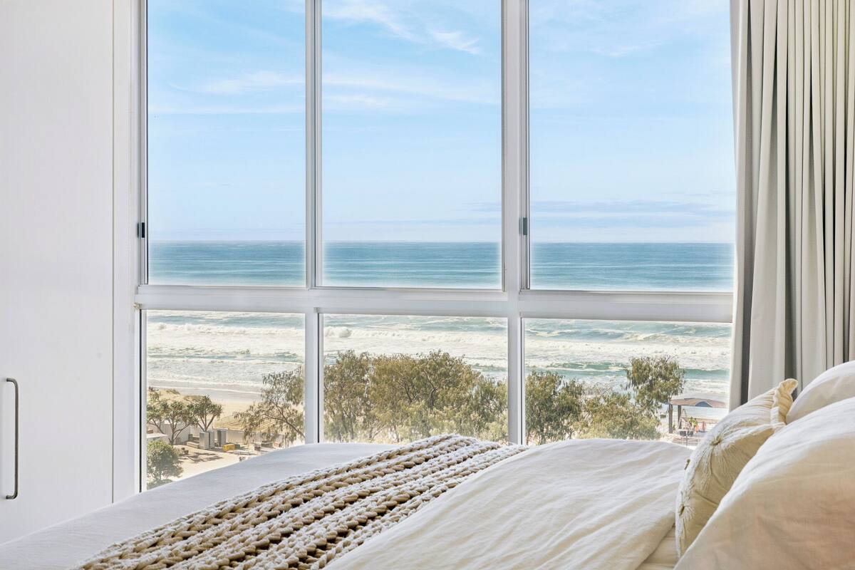 A bedroom with a large window overlooking a beach, a bed with white linens, and a view of the ocean.