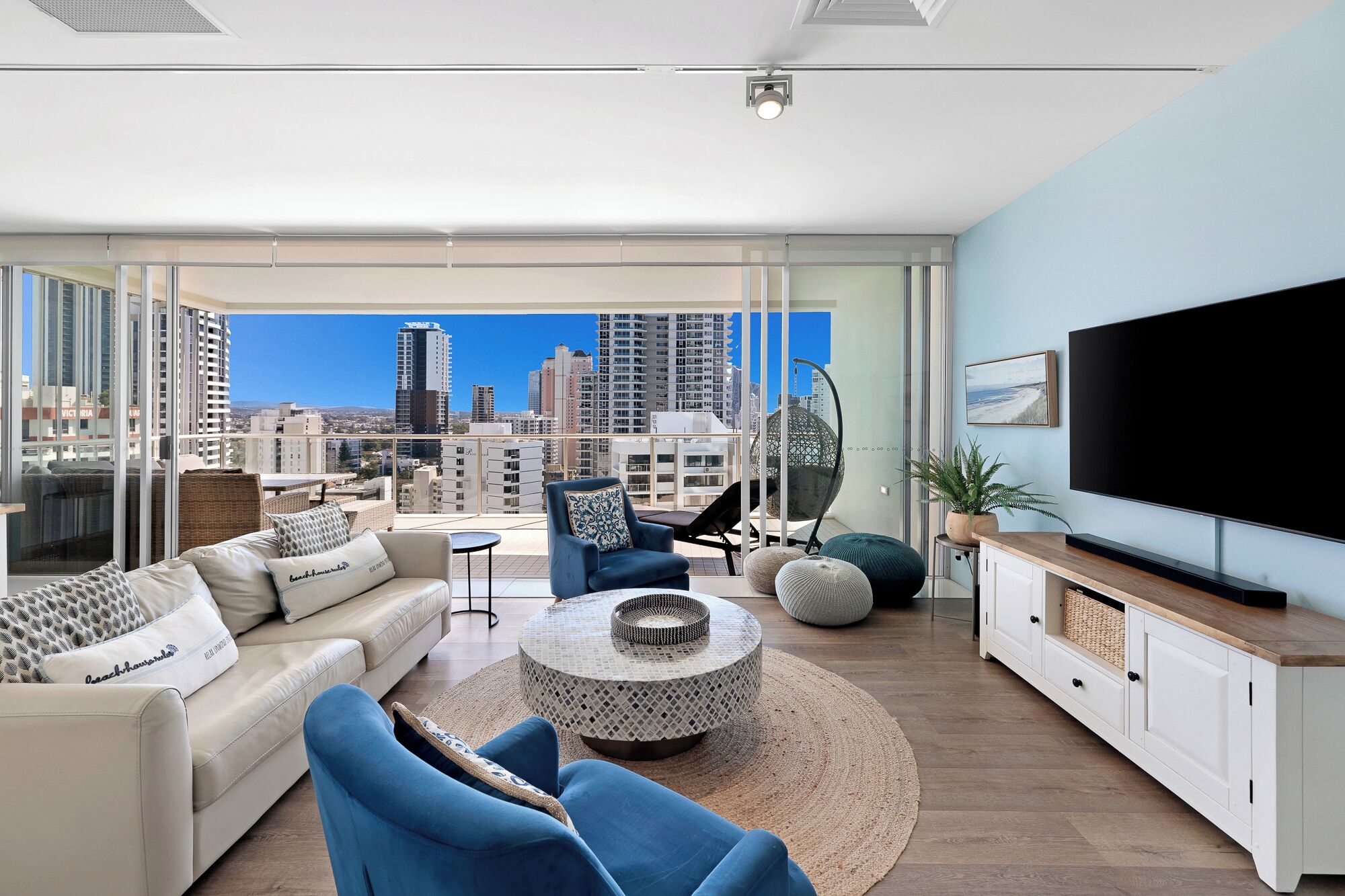 A modern living room with a sofa, chairs, a coffee table, and a TV. The room has large windows with a city view.
