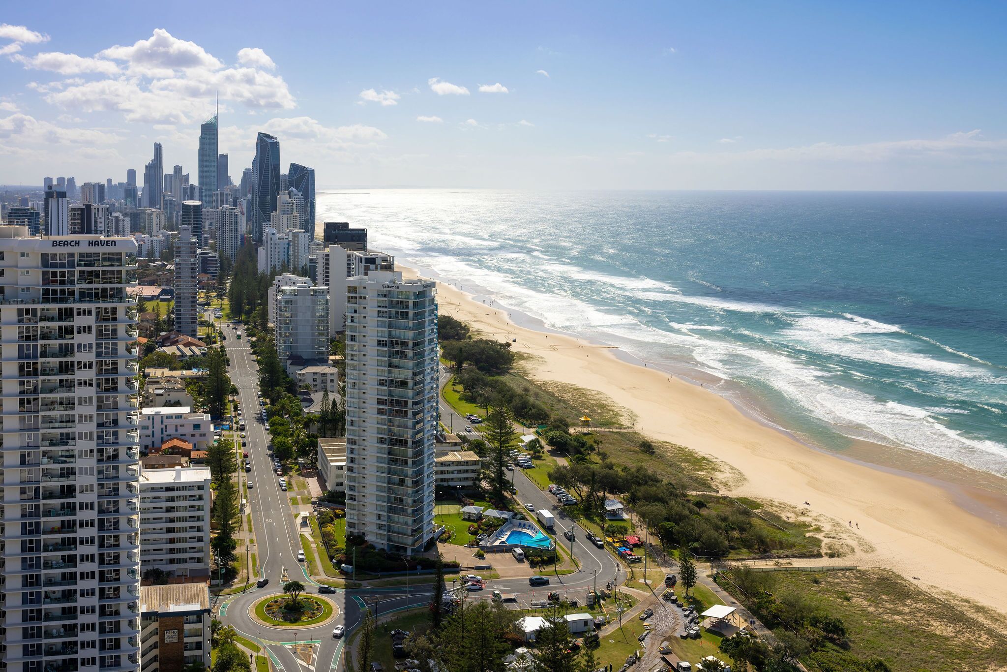 A coastal cityscape with high-rise buildings, a beach, and a road leading to a roundabout.