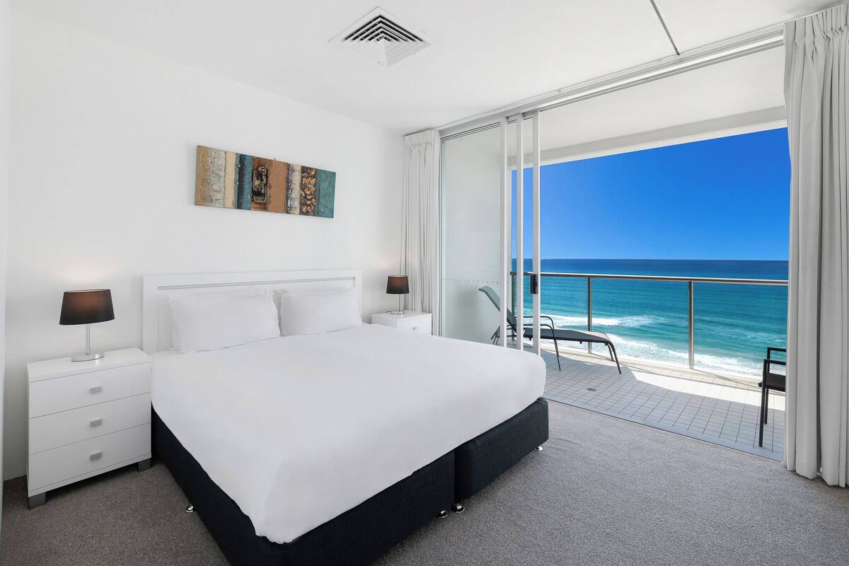 A modern hotel room with a large bed, a painting on the wall, and a balcony with a view of the ocean.