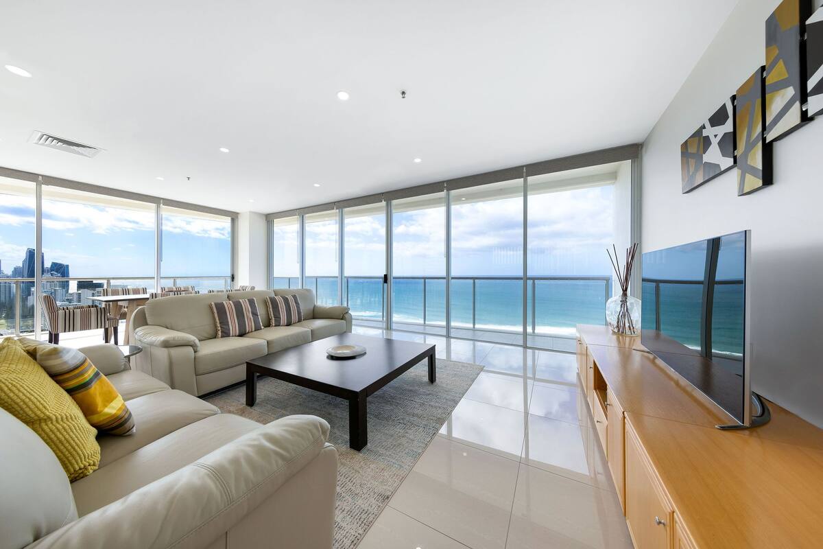 A modern living room with a large window overlooking the ocean, a sofa set, a coffee table, and a flat-screen TV.