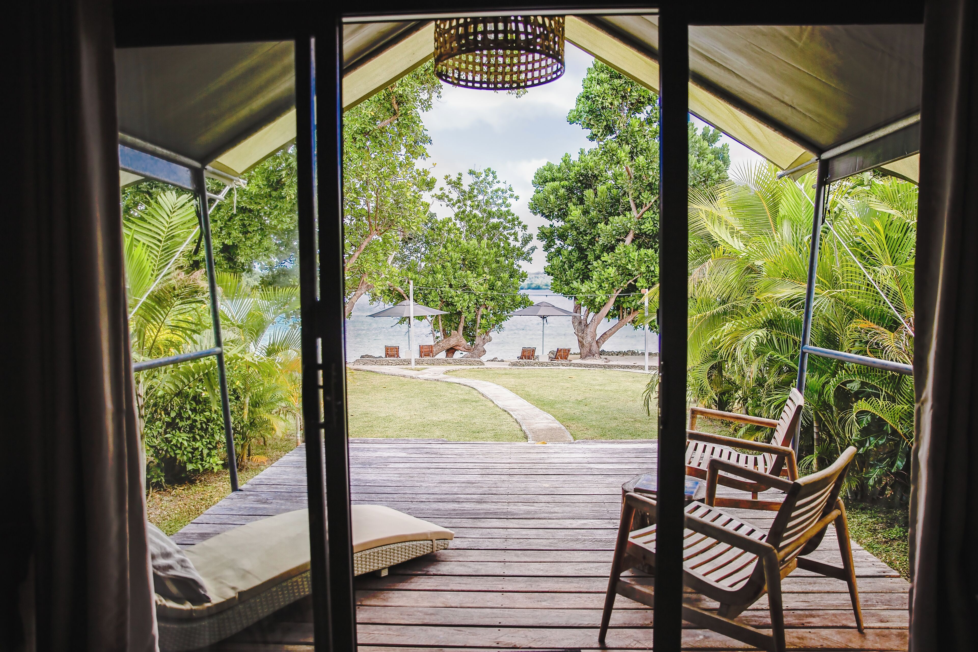 A wooden deck with a bench and a cushion, overlooking a garden with trees and a pathway leading to umbrellas.