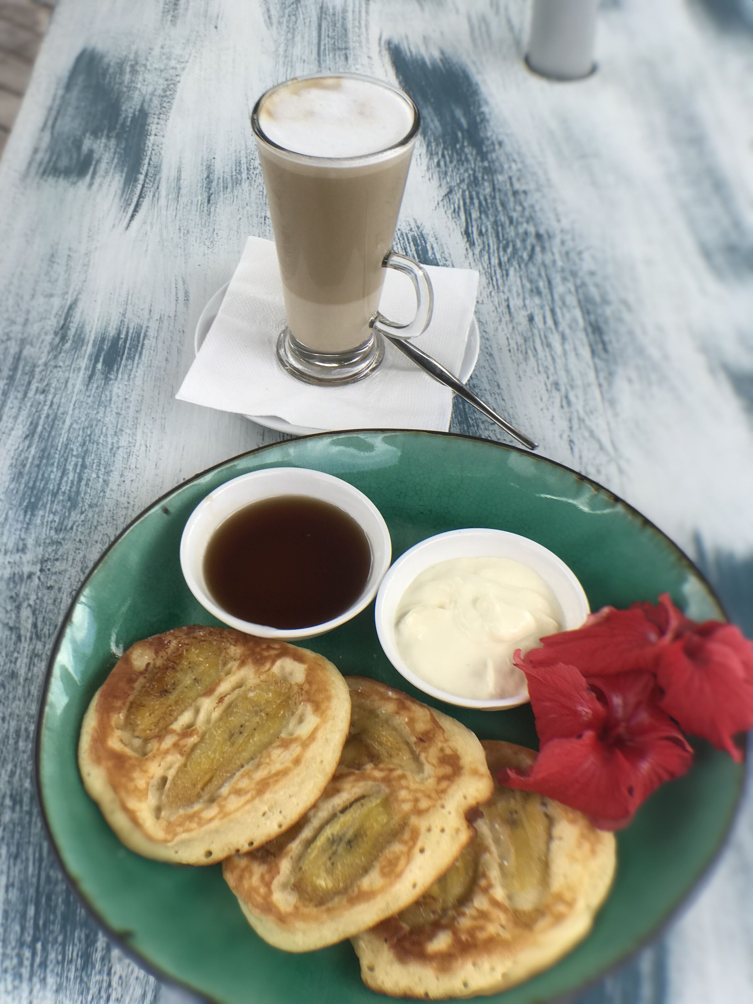 Un plato de panqueques, una taza de café y dos recipientes pequeños con acompañamientos sobre una mesa de madera.