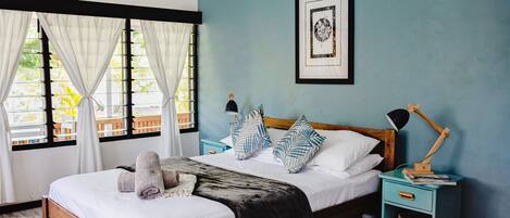 Un dormitorio con cama, cortinas, una mesita de noche con lámpara, un cuadro en la pared y una ventana con vista a palmeras.