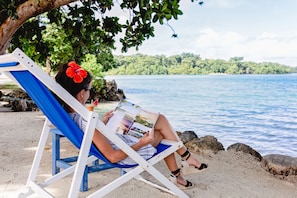 A person sitting on a blue and white beach chair under a tree, reading a magazine.