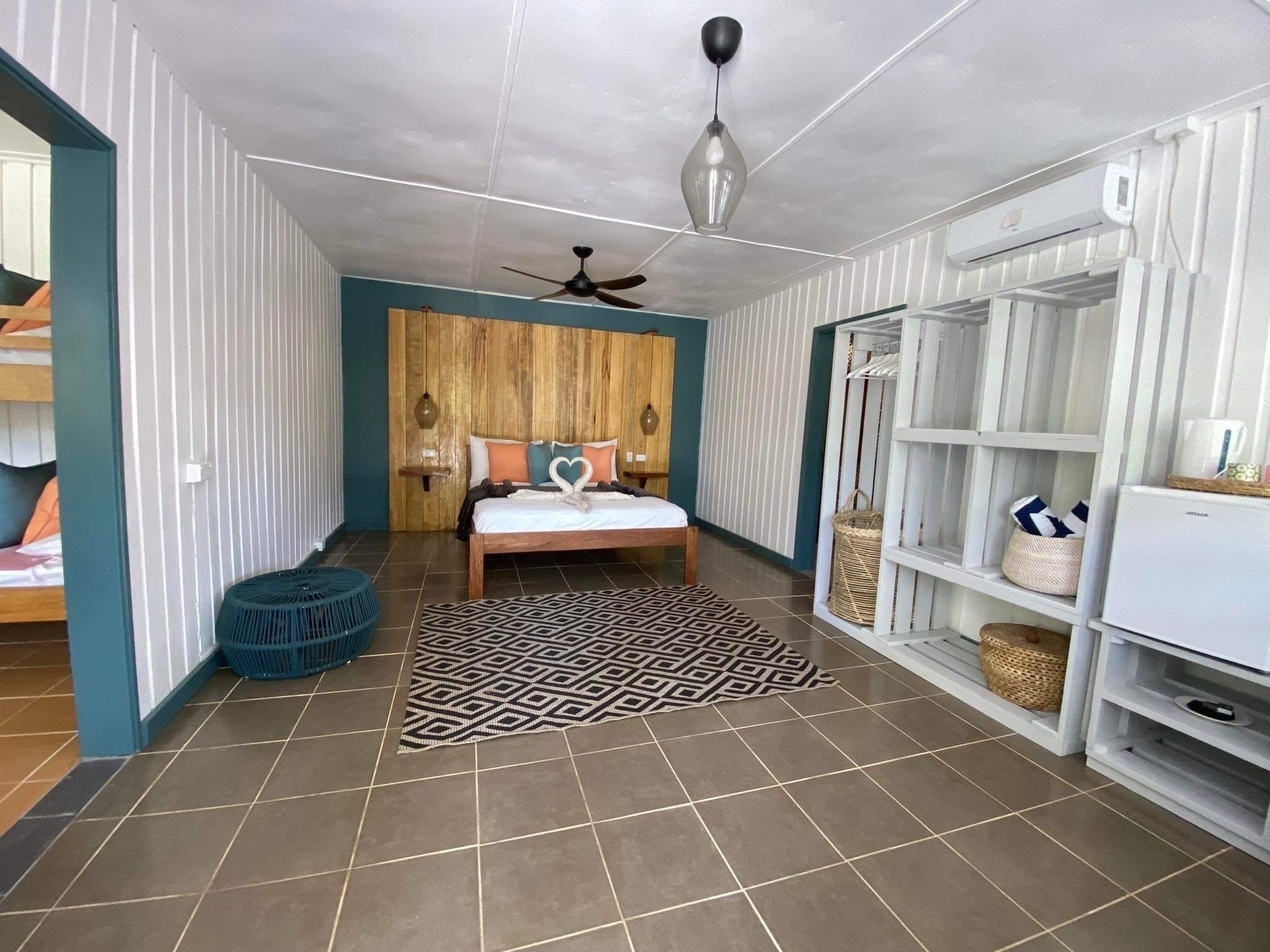 Ein Schlafzimmer mit einem Bett, einem Deckenventilator, einem gemusterten Teppich und einer weißen Regaleinheit mit Körben.