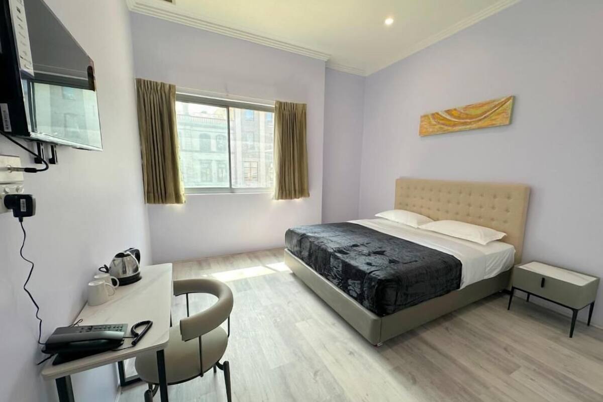 A hotel room with a bed, a desk with a chair, a TV mounted on the wall, and a window with curtains.