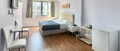 A hotel room with a bed, a desk with a chair, a small table with a kettle, and a refrigerator.