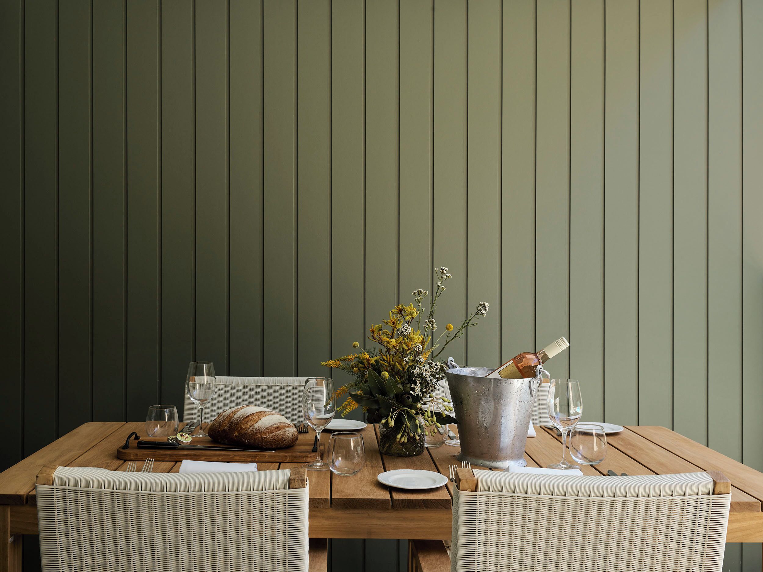 A wooden dining table set with a loaf of bread, wine glasses, a metal bucket, and a wicker chair.