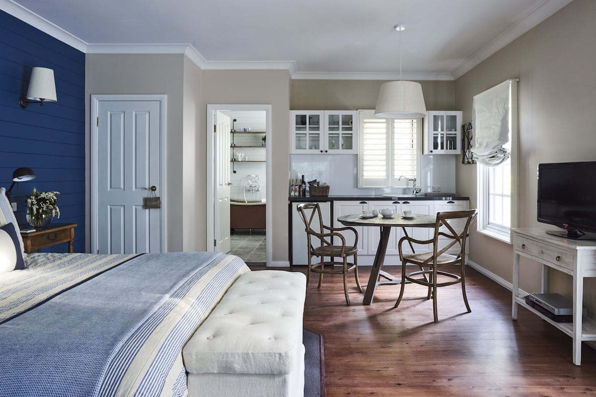 A bedroom with a bed, bedside table, lamp, and a view into the kitchen.