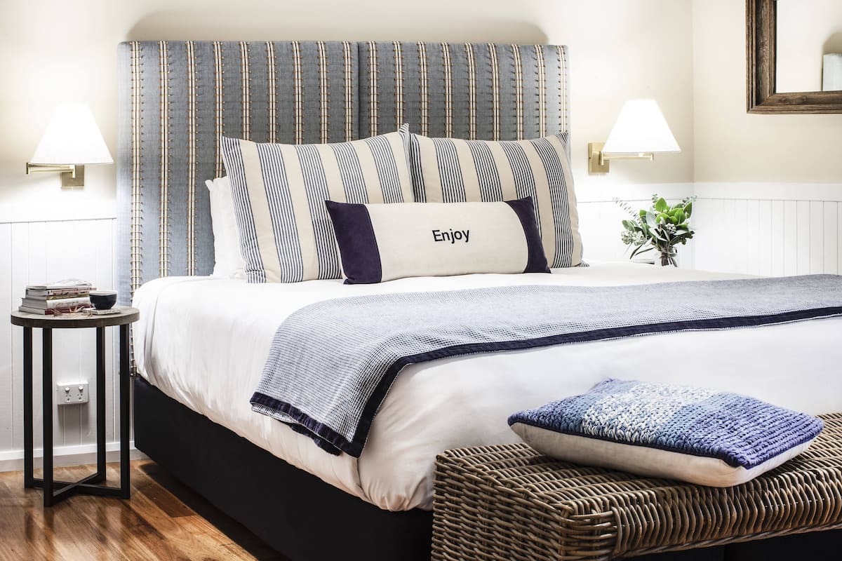 A neatly made bed with striped bedding and a pillow with "Enjoy" text, a small side table with books, a lamp, and a plant.