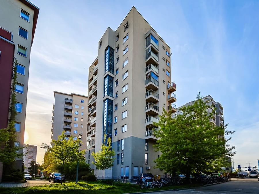 Un edificio residencial alto con varios balcones, rodeado de vegetación y vehículos estacionados.