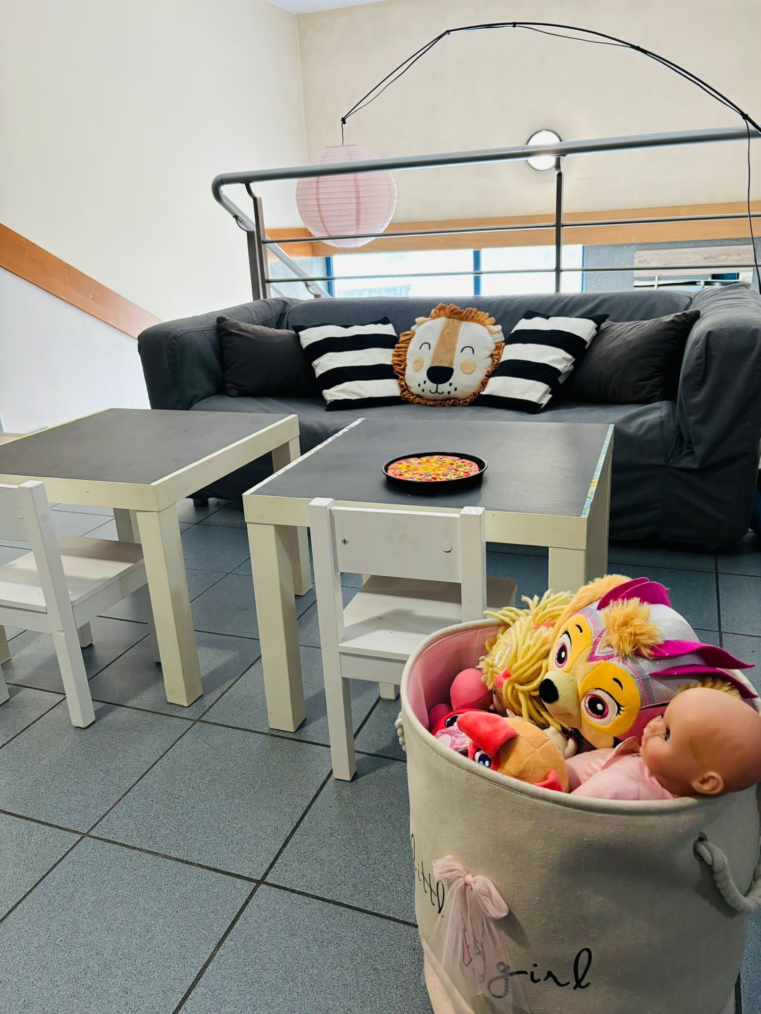 Ein Zimmer mit einer grauen Couch, einem kleinen Tisch, einem Mülleimer mit Spielzeug und einer Pendelleuchte.