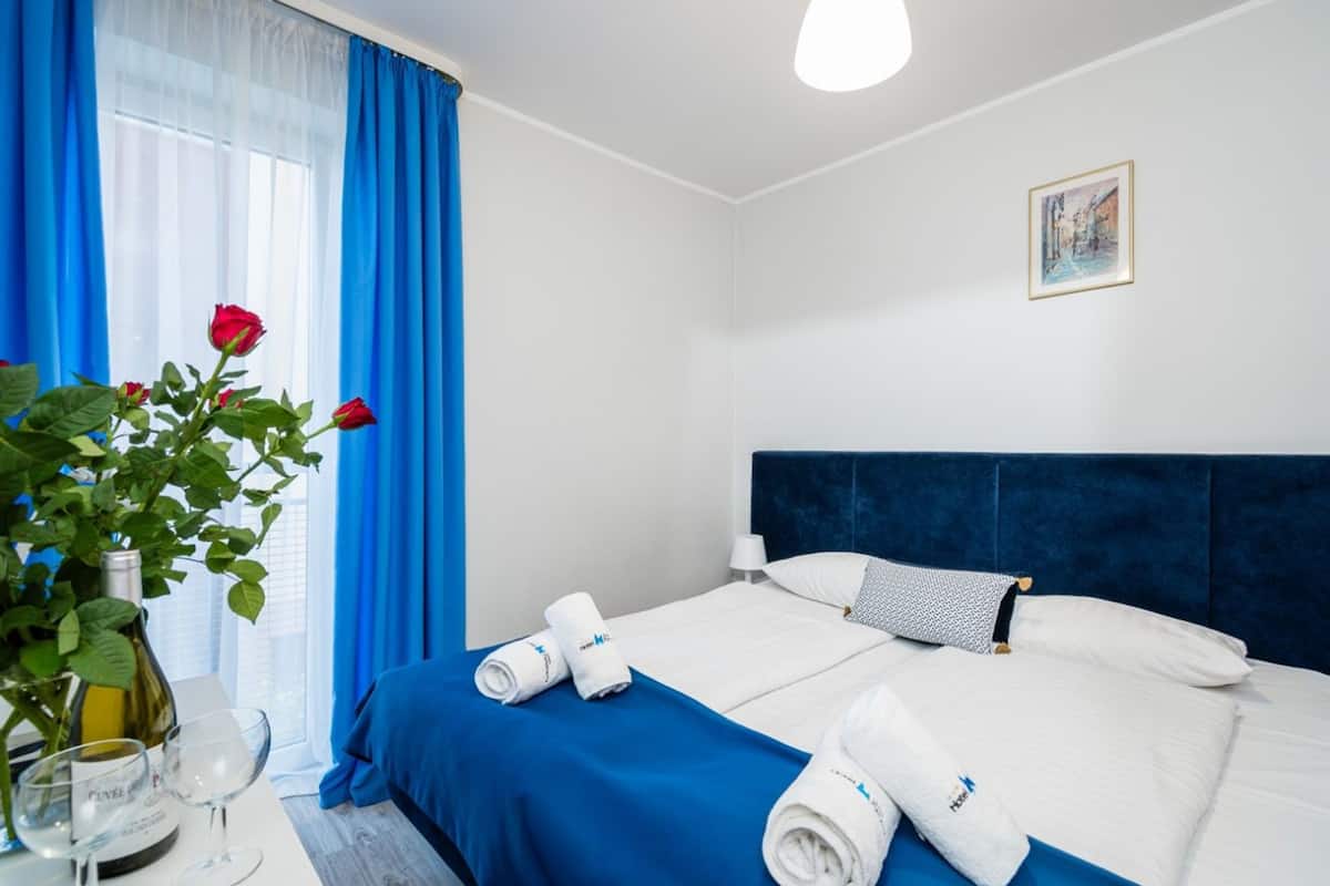 A hotel room with a large bed, blue curtains, a vase with red roses, and a painting on the wall.