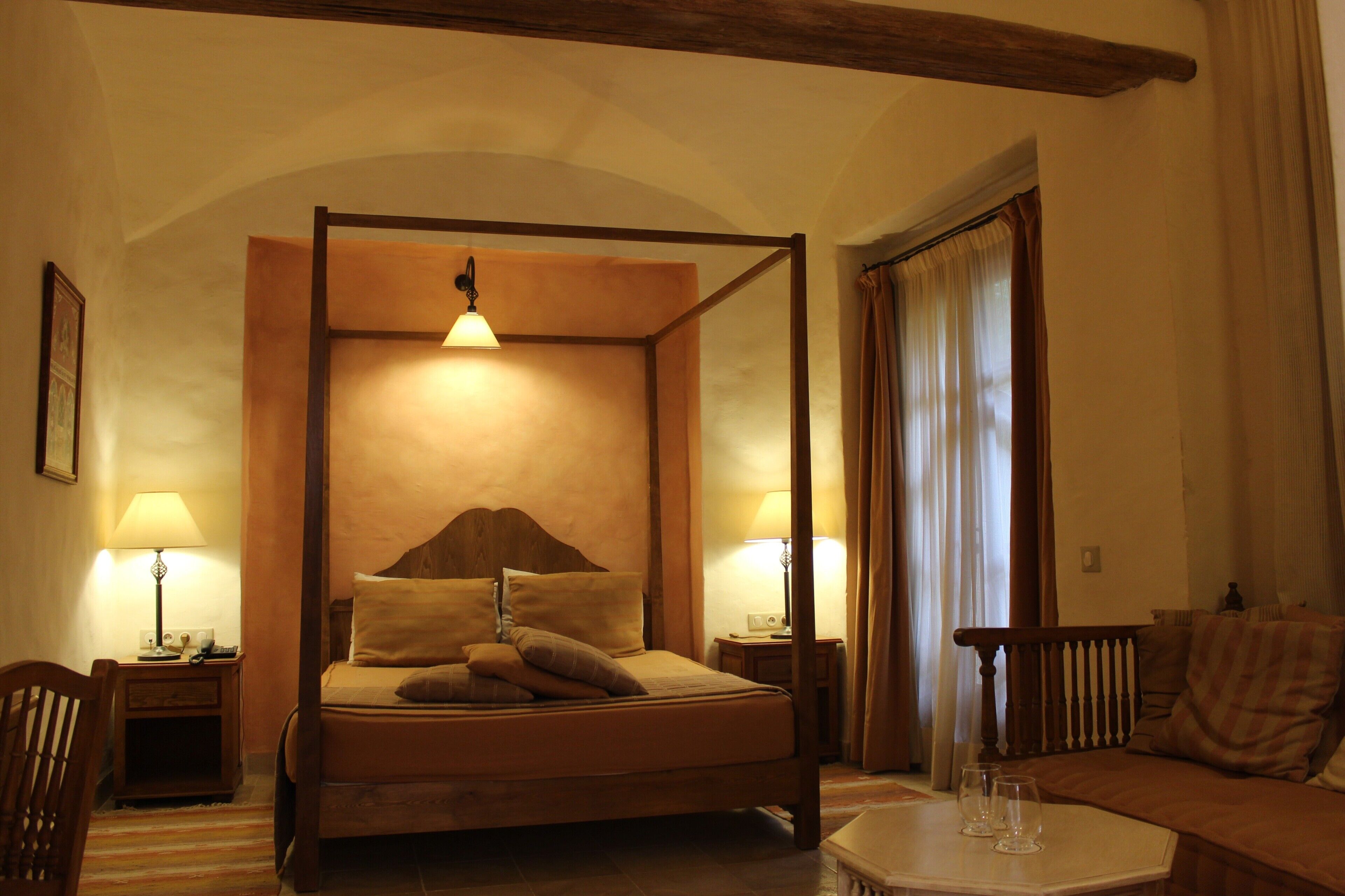 A four-poster bed with a wooden headboard, two bedside lamps, a small table with glasses, and a sofa with a checkered cushion.