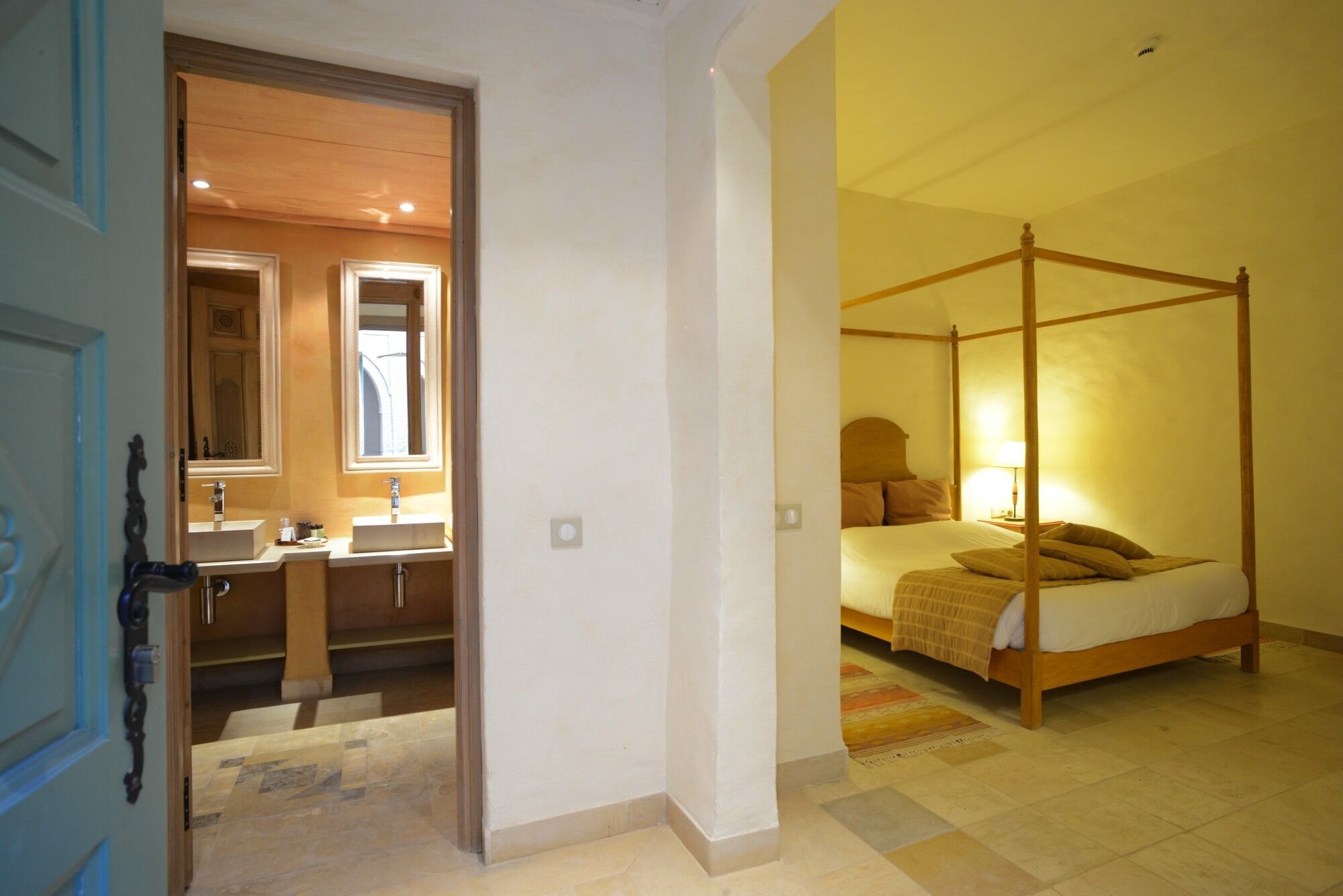 Ein Schlafzimmer mit Himmelbett, Nachttisch mit Lampe und Blick auf ein Badezimmer mit Waschbecken und Spiegel.