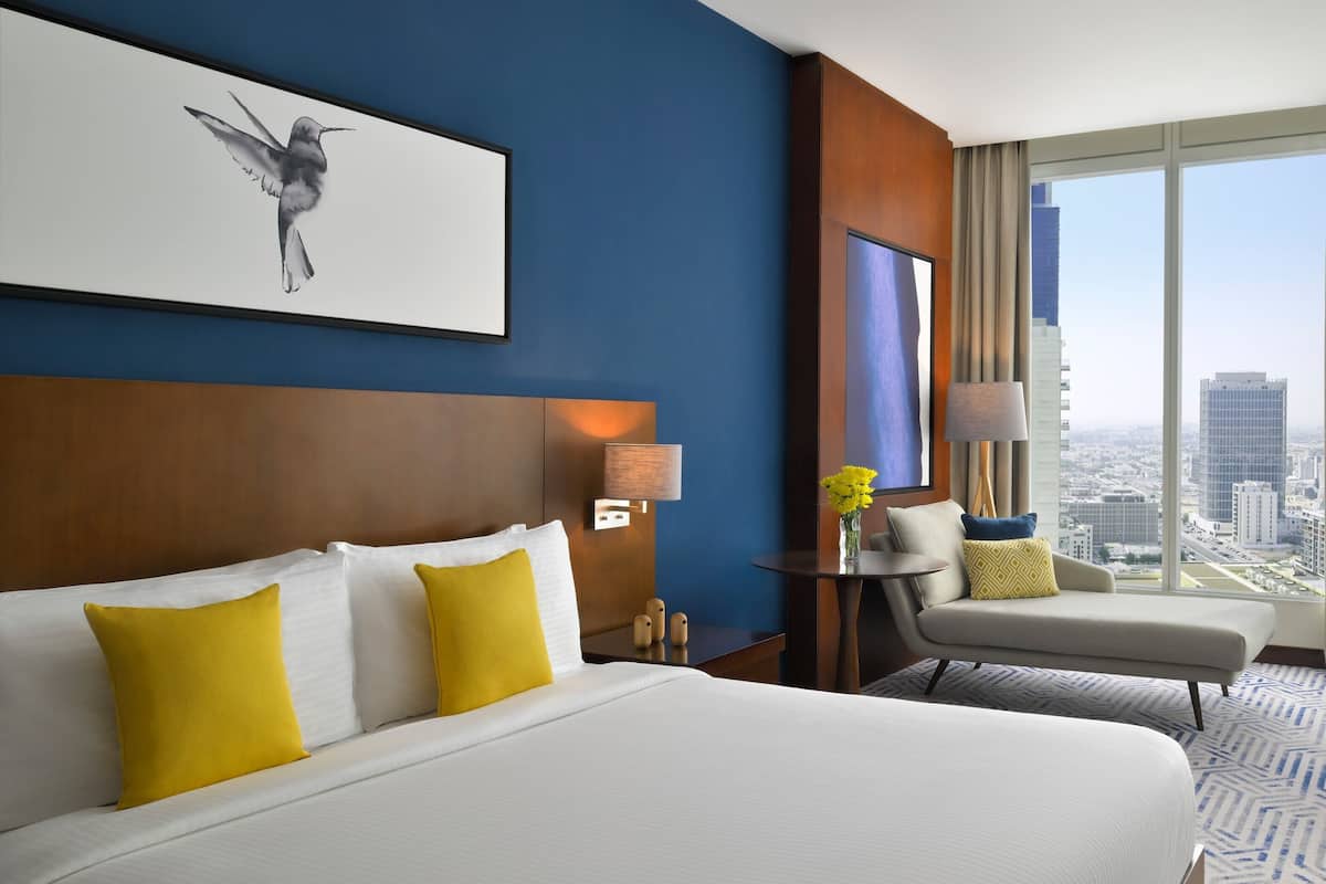 A hotel room with a large bed, a desk, a chair, and a large window with a city view.