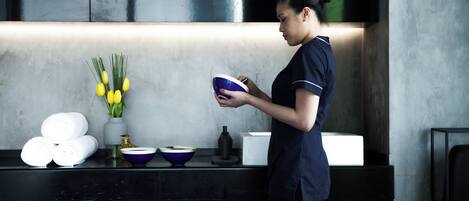A person in a uniform holding a bowl in a modern kitchen with a marble countertop, a vase with yellow flowers, and neatly arranged towels.