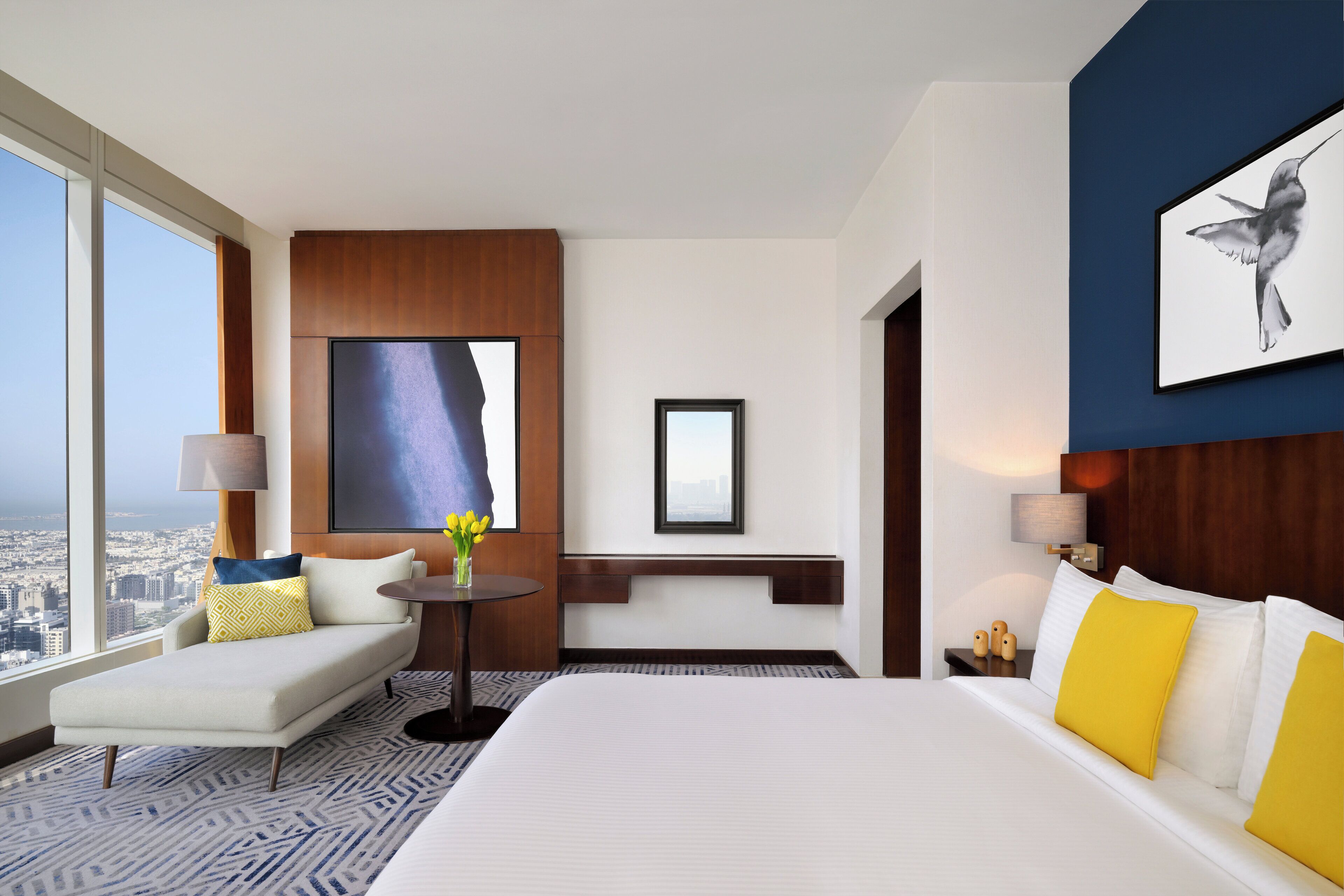 A hotel room with a large bed, a sofa, a small table with flowers, a city view, and a framed picture on the wall.